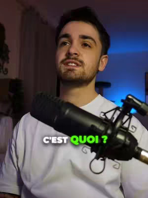 A man with a beard smiles and speaks into a microphone, with a green text overlay that says "C'EST QUOI?" in front. He's wearing a white t-shirt. The background is blurred, with room lighting.
