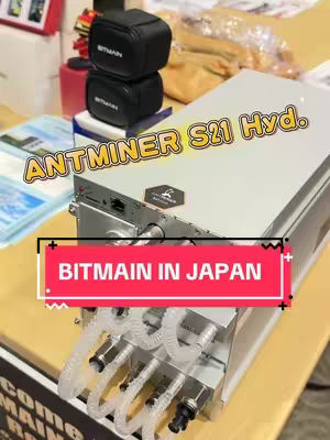 The cover image focuses on the Antminer S21 Hyd, a cryptocurrency mining machine. The machine is silver, prominently displaying the text 'ANTMINER S21 Hyd.' in a yellow outlined font. A bright red banner overlay states 'BITMAIN IN JAPAN.' The composition includes other smaller objects and a blurred background, suggesting an event or exhibition setting. The top left corner shows a small shelf or case with black branded products.