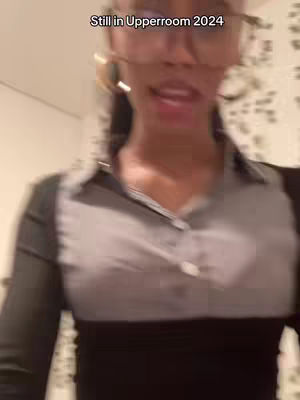 The video shows a woman with glasses and a gray and black top, partially blurred due to the motion and focus. Text overlay reads "Still in Upperroom 2024". The background is light-colored and out of focus.