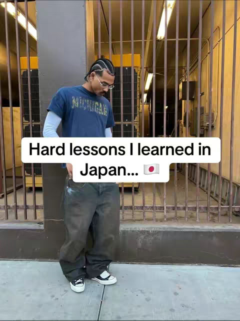 A man wearing a blue shirt and oversized jeans is standing in front of a metal gate, with a text overlay in a white box on the bottom. The background has vertical metal bars. The text overlay has the text : "Hard lessons I learned in Japan..." and the Japanese flag.