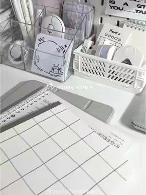 The image showcases a clean, well-organized desk setup. A clear plastic organizer holds pens and pencils. A small white basket and two transparent containers are filled with various office supplies. A notepad with a playful alien design and grid-lined papers are visible, with a predominantly white and pastel color palette, fostering a calm and study-friendly environment.