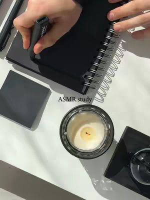 A top-down shot showing a white surface with study materials. There's a black notebook with a spiral binding, with a hand holding a black marker over the page, as if writing. To the side lies a closed black square, a jar candle, and parts of a black pen.