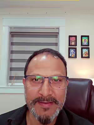 A man with glasses and a mustache looks directly at the camera. The background includes a window with blinds, decorative frames, and wall details. The colors are muted neutrals.