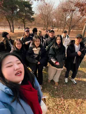 A group of people, mostly young adults, are gathered outdoors on a sunny day. A woman in the foreground is taking a selfie, smiling with her mouth open and her face positioned slightly off-center. She is wearing a blue jacket and a red scarf. Behind her, others in varying styles of winter clothing stand, some looking at the camera, others away. The background includes trees and grass, suggesting a park or outdoor setting.
