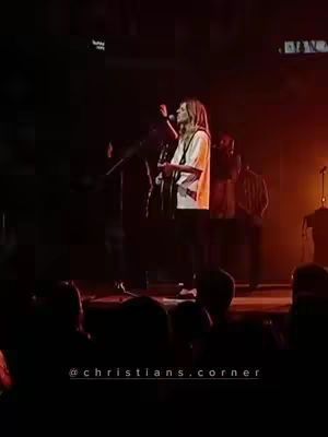 A stage performance with a female singer playing a guitar in front of a crowd. The image is dark with spotlight effects highlighting the singer and the stage.