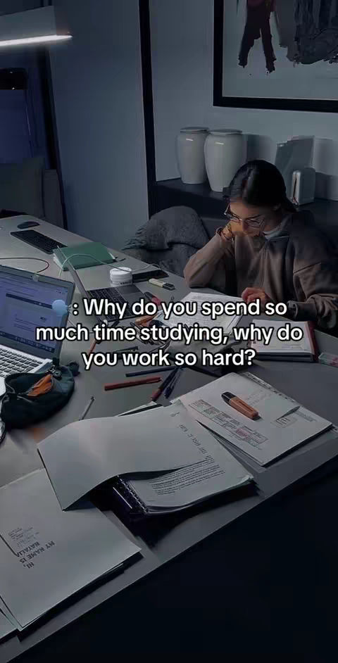A dark, interior shot of a person studying at a desk. The  person, wearing glasses and a dark-colored top, is leaning over a desk covered in papers, a laptop and various stationery. A light filters in from a window or a source above, illuminating the workspace. The text overlay asks “Why do you spend so much time studying, why do you work so hard?”