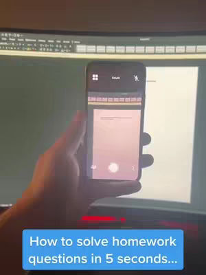 A person's hand is holding a smartphone, displaying an app interface with a document-like appearance. In the background, there is a dark monitor and a wall.
