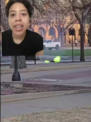 A woman with short, dark hair is centered in the frame. She is wearing a black t-shirt and appears to be speaking. Behind her, a blurred outdoor scene includes trees, an arched brick building, and a white truck. A greenish-yellow burst of light is visible on the ground.