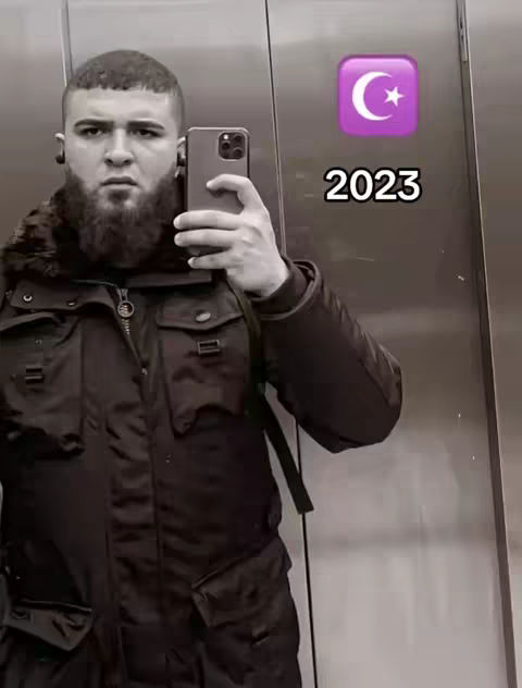 A man holding a phone, taking a selfie in an elevator. The background is the inside of the elevator, which is made of a metallic material. On the upper right corner is a symbol with a purple background, and the number 2023 is below it. The man is wearing black jacket. The man has a beard