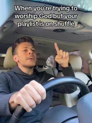 A man is sitting in the driver's seat of a car, looking up with a slight smile and pointing his index finger up. Text overlayed on the image says 'When you're trying to worship God but your playlist is on shuffle'. The car's interior is visible with beige seats and the top of the car.
