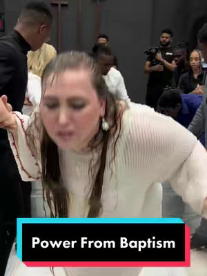 The image captures a moment of baptism, with a woman as the central figure. She is in a white dress, leaning forward with hair wet as if just emerged from water. Several people surround her, some holding her, while others stand behind and to the sides, including possible cameramen in the background. The overall setting suggests a church environment, with a cross visible and people dressed in formal attire. The bottom of the visual has an added text saying "Power From Baptism".