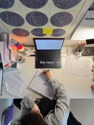 An overhead shot of a study desk with a laptop, papers, pens, and a person writing on paper. A constellation map is taped to the wall. The colors are muted with a focus on the desk and the person's activity.