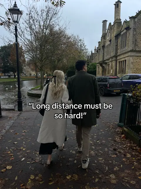 A couple walks down a paved path, seen from behind. The woman, wearing a white coat and a black dress, holds the right hand of the man, who has a green jacket on. They are surrounded by a blend of architectural buildings with what seems to be a pond or river to the left.