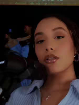 A selfie-style shot focuses on a woman's face, with the background blurred, suggesting an inside setting. The woman has light brown skin, and the features are clear. The layout is a close-up, emphasizing the subject. She's wearing a light-blue collared shirt with a silver necklace.