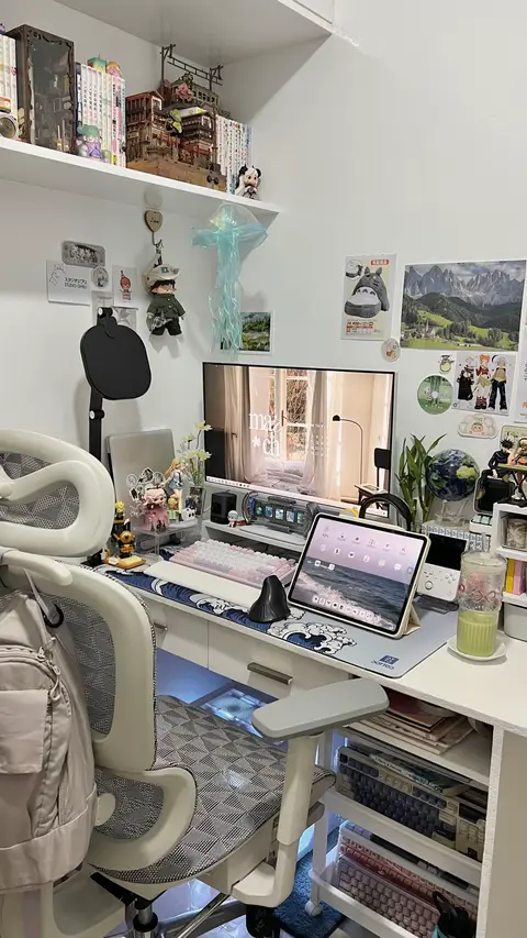 A well-lit room with a neatly organized desk. A large monitor displaying a cozy interior scene. A laptop and an iPad are visible on the desk, surrounded by various decorations like toys, plants, and framed pictures. A white and gray ergonomic chair is in front of the desk. Shelves above the desk are filled with books and decorative items.
