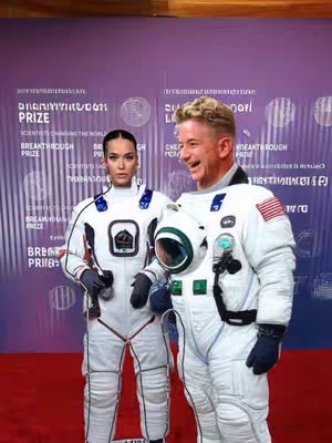 The image features Katy Perry and a man, likely Jeff Bezos, in what appears to be an astronaut-themed setting with event branding in the background. They are on a red carpet, both wearing white astronaut suits. Katy Perry has her hair pulled back and is wearing a serious expression, while the man is smiling as he holds an astronaut's helmet. Text in the background is partially visible, suggesting a science or award event.