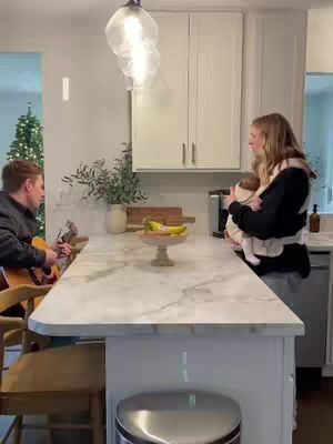 A family worship scene takes place in a modern kitchen. A man is playing the guitar, while a woman holds a baby in a carrier. The kitchen has white cabinets, a marble countertop island, and a Christmas tree visible in the background.