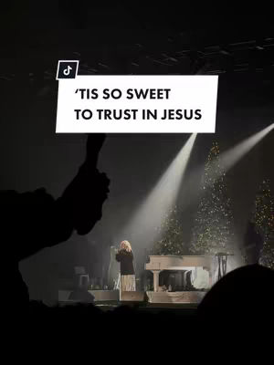 The image shows a concert setting with a silhouetted hand holding a sign that reads "'Tis so sweet to trust in Jesus." In the background, a woman is on stage singing near a white piano with Christmas trees on either side. Bright spotlights shine down.