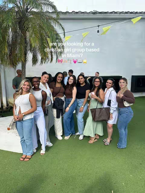 A group of young women smiling and posing outdoors on a sunny day. They are standing on green grass, and a large palm tree is visible on the left. Behind them, there's a white wall and some decorations. The overall vibe is positive and friendly.