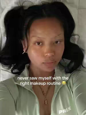 A selfie-style shot shows a young woman lying on white bedding, looking directly at the camera with a soft, possibly makeup-enhanced, appearance. Her dark hair is styled into two pigtails. She wears a light green zip-up top and a delicate necklace. The image has a soft, natural lighting.