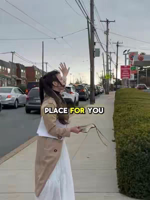 A woman in a white dress and tan coat stands on a sidewalk next to a street with cars. She is looking up and raising her arm, with a sign that includes the words "PLACE FOR YOU" in yellow. The sky is overcast.