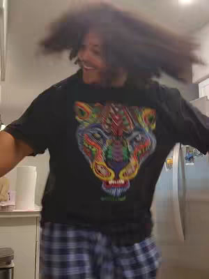 A man with long, dark, curly hair is captured at eye level, mid-motion, likely in the midst of a dance or energetic movement. The lighting appears natural and even, highlighting the details of his outfit: a black t-shirt adorned with a vibrant, colorful graphic of a mythical creature's head and patterned pajama pants in shades of blue and white, and he is smiling as he moves within the room. The background is blurred, with hints of a kitchen setup.