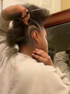 A person, likely a woman, is seen from the shoulders up styling dry, textured hair into cornrows. The hands are holding a comb and working on the hair. The person is wearing a light-colored, long-sleeved shirt. A mirror is visible in the background, reflecting more of the person and the room. The image has a warm, indoor lighting feel.