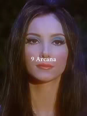 A close-up of a woman's face with dramatic makeup, including bold blue eyeshadow and bright pink blush. The text '9 Arcana' is superimposed in white at the bottom-center. The background is out of focus, with a dark, possibly forested setting.