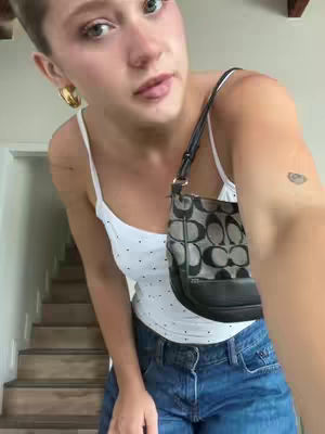 A young woman stands indoors, slightly angled towards the camera. She wears a white tank top with small black polka dots, paired with blue denim jeans. A small black Coach purse hangs from her shoulder across her body. Her short hair is styled back, and gold hoop earrings adorn her ears. The background reveals a staircase and a glimpse of a white wall, suggesting a home interior.