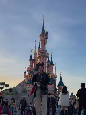 The image shows a woman standing in front of the Sleeping Beauty castle at Disneyland Paris. She wears Mickey Mouse ears. The castle is in the background under a light blue sky, and the people are walking around in the foreground.