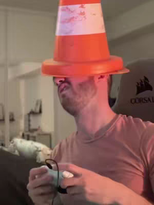A man wearing an orange traffic cone on his head is playing a video game. He is holding a game controller and looking down at the screen. The background includes a gaming chair and some blurred-out objects.
