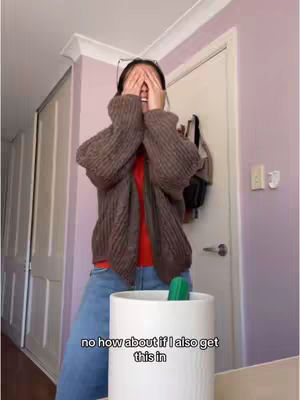 The cover image shows a person with their hands covering their eyes. They are standing in a room with a white door and neutral-colored walls. The person is wearing a brown sweater over a red shirt, with blue jeans. A green object is positioned in a white pot, which sits in the foreground of the image.