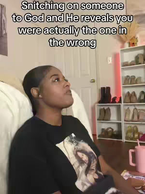 A woman with a dark complexion and short hair is seated indoors, gazing upwards with a contemplative expression. The background features a door, a bookshelf lined with various items, and a pink cup with a straw. The lighting seems natural, illuminating the scene from one side.