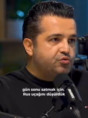 A man in a black shirt is speaking into a microphone. Text overlayed near the bottom of the frame says 'gün sonu satmak için, Rus uçağını düşürdük'. The background is blurred, suggesting an indoor setting with a microphone in the foreground.