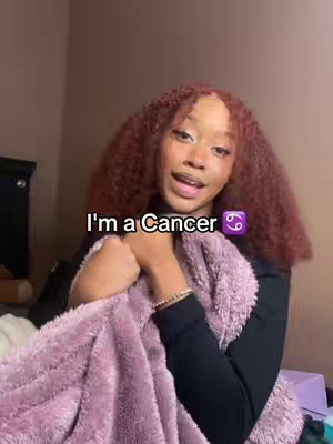 A young woman with curly red hair is centered in the frame, holding a pink fuzzy blanket around her. The text overlay reads 'I'm a Cancer' with a Cancer zodiac symbol. The background is a simple neutral wall.