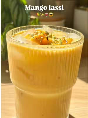 A close-up shot of a glass filled with a creamy, yellow-orange beverage, likely a mango lassi, garnished with small pieces of what appears to be mango and nuts. Ice cubes are visible in the drink. The glass is set against a softly blurred background, with hints of green foliage and possibly another surface reflecting warm sunlight. The glass has a ribbed design.