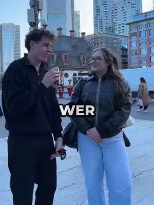 A medium shot shows a man and a woman in an outdoor setting. The man, on the left, is wearing a black jacket and dark pants, holding a clear cup up. The woman, to his right, wears glasses and light-colored pants with a black leather jacket, smiling at the man. The background features city buildings and a pedestrian area.
