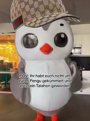 The image shows a person in a penguin costume wearing a Gucci hat. The penguin is the central figure, with a white body, grey wings, and orange feet. The background appears to be a room with a kitchen counter or shelves visible.