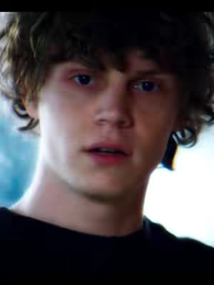 A close-up shot focuses on the face of Evan Peters, who has curly dark hair. His eyes are blue and he appears to be looking slightly off-camera with a subtle expression. He is wearing a black shirt and the background is slightly blurred, having a blue hue.