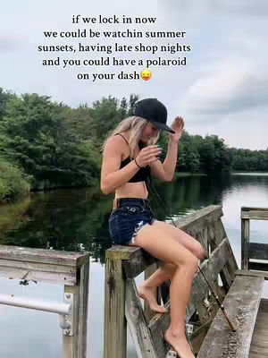 A woman in a black cap and crop top sits on a wooden structure at a lake or river. Text is added to the top of the photo.