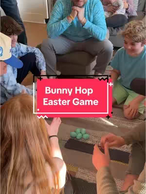 A group of people are seated in a circle on a patterned rug. A man is sitting on a small cube, and a group of children are gathered around. In the center, there are green eggs and a sign over that reading "Bunny Hop Easter Game".