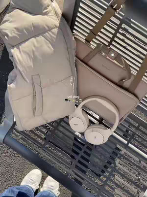 The image shows an overhead view of a beige puffer vest, a light-colored handbag and matching laptop case, and a pair of white headphones lying on a metallic surface, with the feet of a person wearing white sneakers visible at the bottom.
