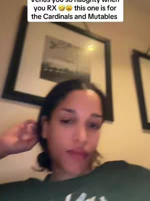A woman with dark hair is centered in the frame from the chest up. She is wearing a dark green top resting her right hand near the side of her head. Behind her are framed pictures and tan wall. There is text in the top left corner of the image.