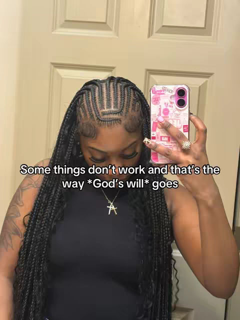 A selfie-style image featuring a Black woman with long, braided hair and a black top. She is holding a pink and white phone, revealing part of her face and a cross pendant necklace. The background is a light-colored door, and text overlayed on the image.