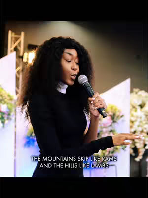 A woman with long, dark hair is centered, holding a microphone and singing in front of a soft, blurred background with flower-like decorations. The woman is wearing a black long-sleeved top over a white undershirt. Text at the bottom of the screen quotes biblical verse.