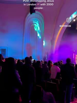 Image captures a worship service inside a large venue with high ceilings and arched features. The foreground is filled with the backs of people's heads, suggesting a crowded space. Light streams down, creating columns of illumination, with purple and blue hues illuminating the back. Text overlays at the top provide context, and a social media handle is present, indicating potential branding.