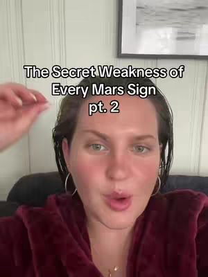 A woman with fair skin and short brown hair is centered in the frame. She has large silver hoop earrings and is wearing a burgundy robe. The text overlay reads, 'The Secret Weakness of Every Mars Sign pt. 2'. The background features a framed piece of art.