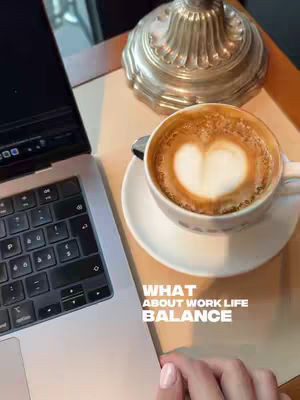 The image shows a top-down view of a workspace. A laptop is on the left, coffee with a heart-shaped latte art in the center, a decorative lamp is on the far right and a female hand with pale pink nail polish is visible in the bottom right corner. Text overlayed on the image reads, "WHAT ABOUT WORK LIFE BALANCE."
