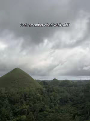 A wide shot of a scenic landscape under a cloudy sky. The foreground is dominated by rolling green hills covered in trees. The sky is overcast with shades of gray. In the center is text, overlayed onto the sky.