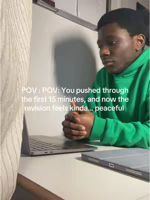 The cover image shows a young Black man in a green hoodie, sitting at a desk with a laptop computer open in front of him and a tablet next to it. He is looking down and to the side pensively with his hands clasped in front of him. The scene appears to be a home office or study space, with a whiteboard and a partially visible patterned curtain in the background. Text overlayed on the image reads, "POV: You pushed through the first 15 minutes, and now the revision feels kinda... peaceful."